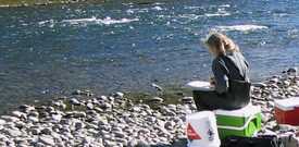 Femme assise sur une glacière à côté d’une rivière, enregistrant les résultats de la qualité des eaux sur une fiche de terrain