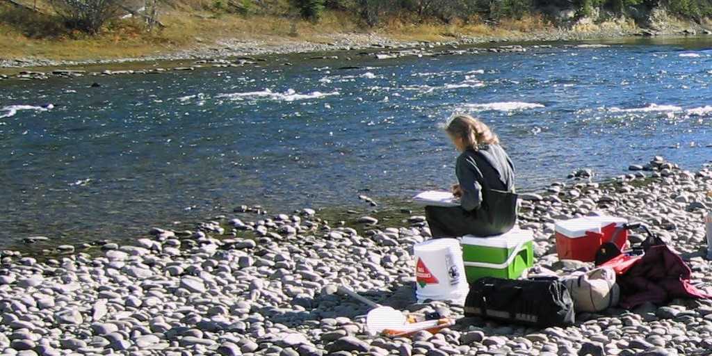 Woman sitting on a cooler next to a river recording water quality results onto a field sheet