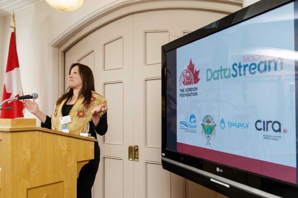 Lana Lowe at a podium next to a screen displaying logos for the gordon foundation, mackenzie datastream, the government of the northwest territories, Fortn Nelson First Nation, Tesera, and CIRA
