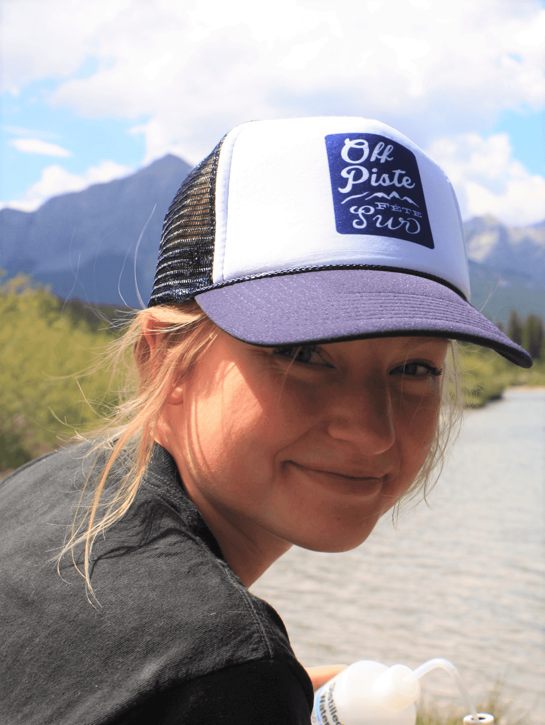 Mary Kruk, Water Data Specialist, smiling with a blue and white cap and mountain and stream in the background.
