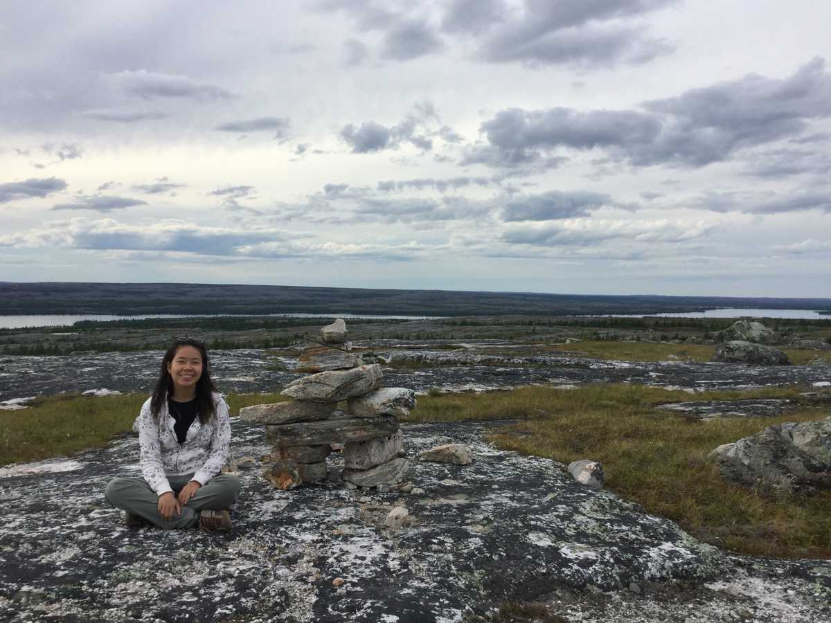 Lilian sitting beside an inukshuk with a view of the tundra and river in the background --- Lilian assise à côté d'un inukshuk avec vue sur la toundra et la rivière en arrière-plan